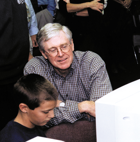 An intrigued Charles Koch looks over the shoulder of a young student working at a computer in the foreground.