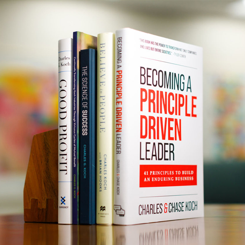A stack of five books authored by Charles Koch on a wooden desk, with a blurred background.
