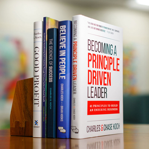 A stack of five books authored by Charles Koch on a wooden desk, with a blurred background.